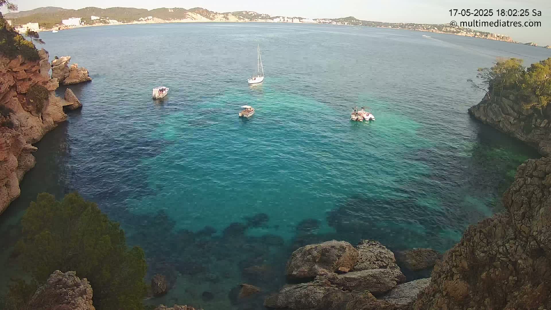 A calm, clear turquoise ocean cove is surrounded by rocky cliffs and several small boats are visible in the water, with a distant shoreline in the background.