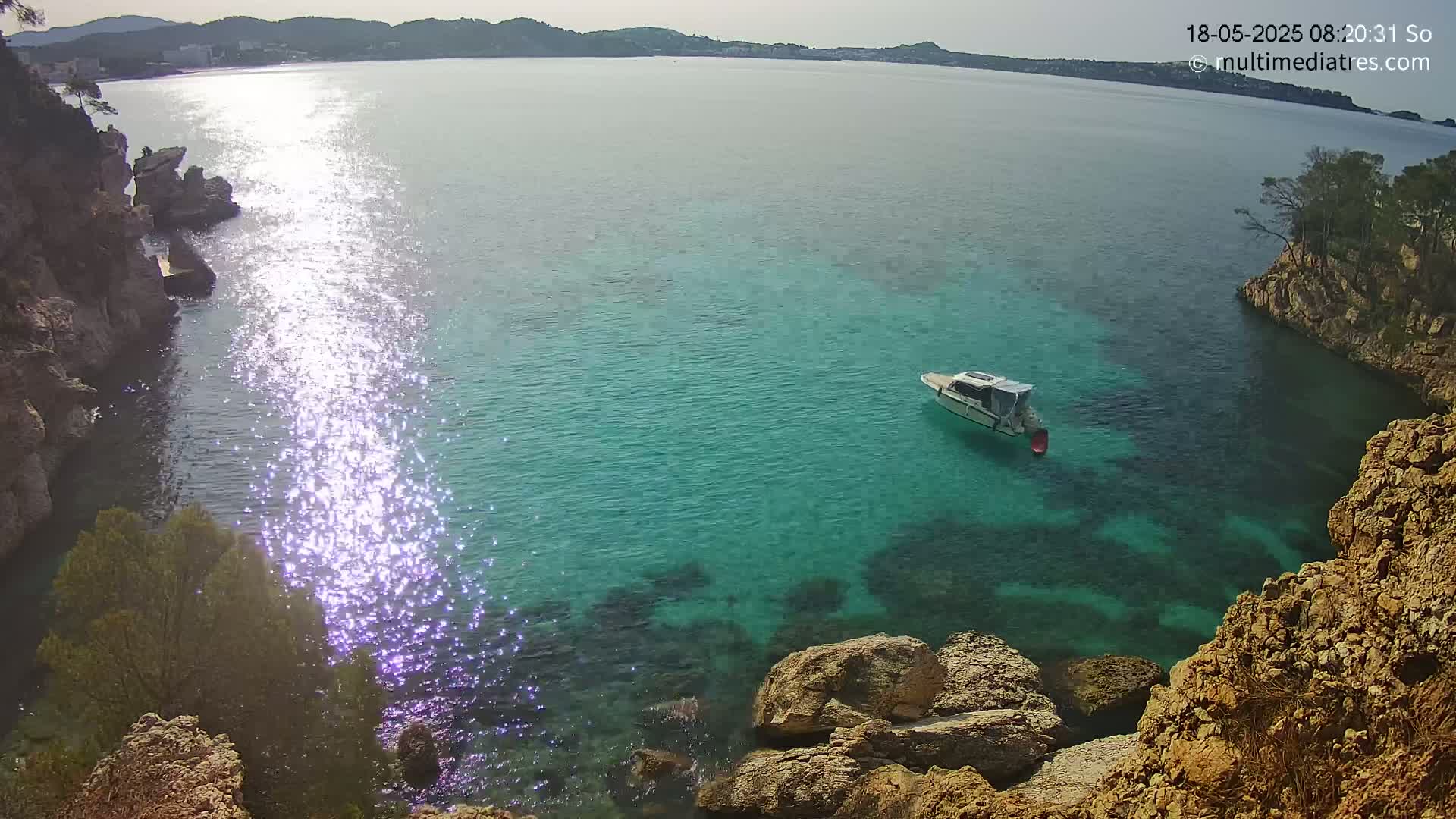 A small boat sits in a calm, clear turquoise cove surrounded by rocky cliffs under a bright, sunny sky.