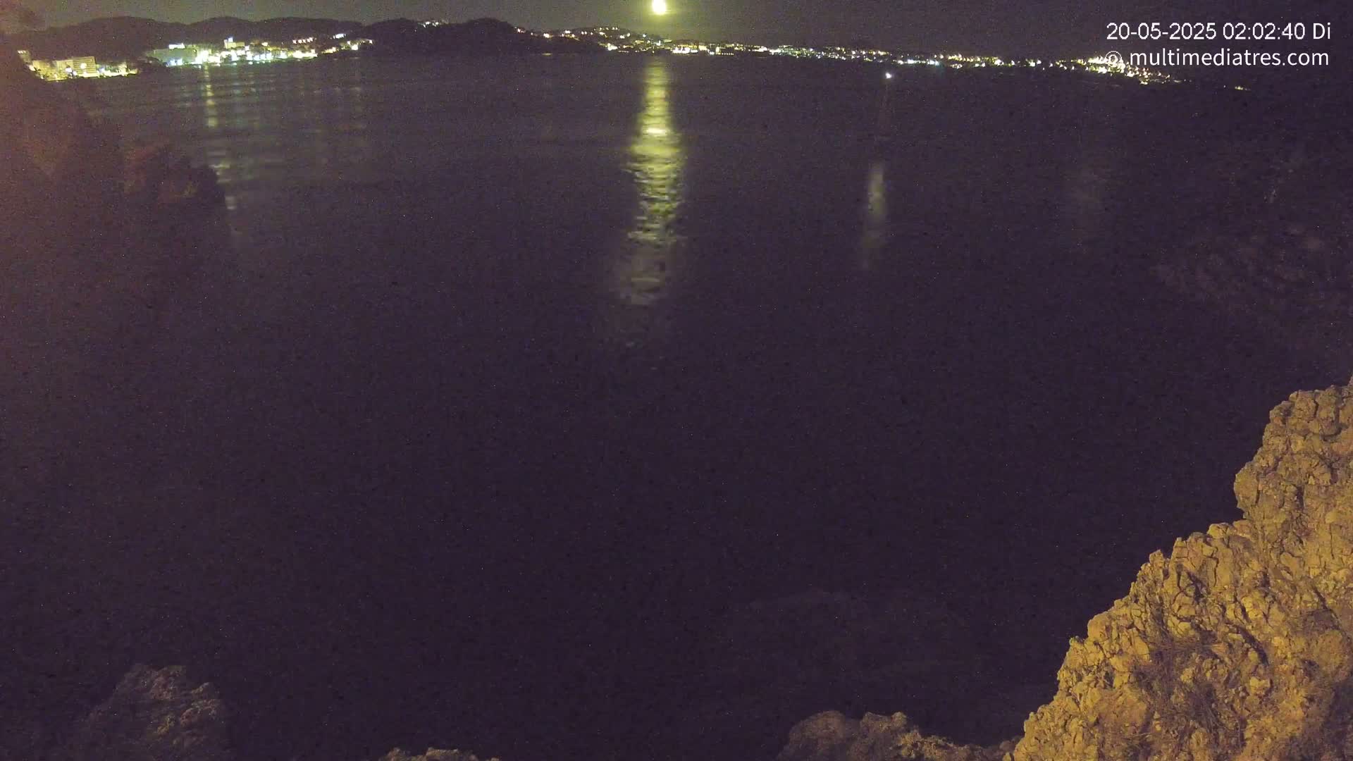 A nighttime view of a moonlit body of water with a distant town's lights visible in the background, partially obscured by a rocky foreground.