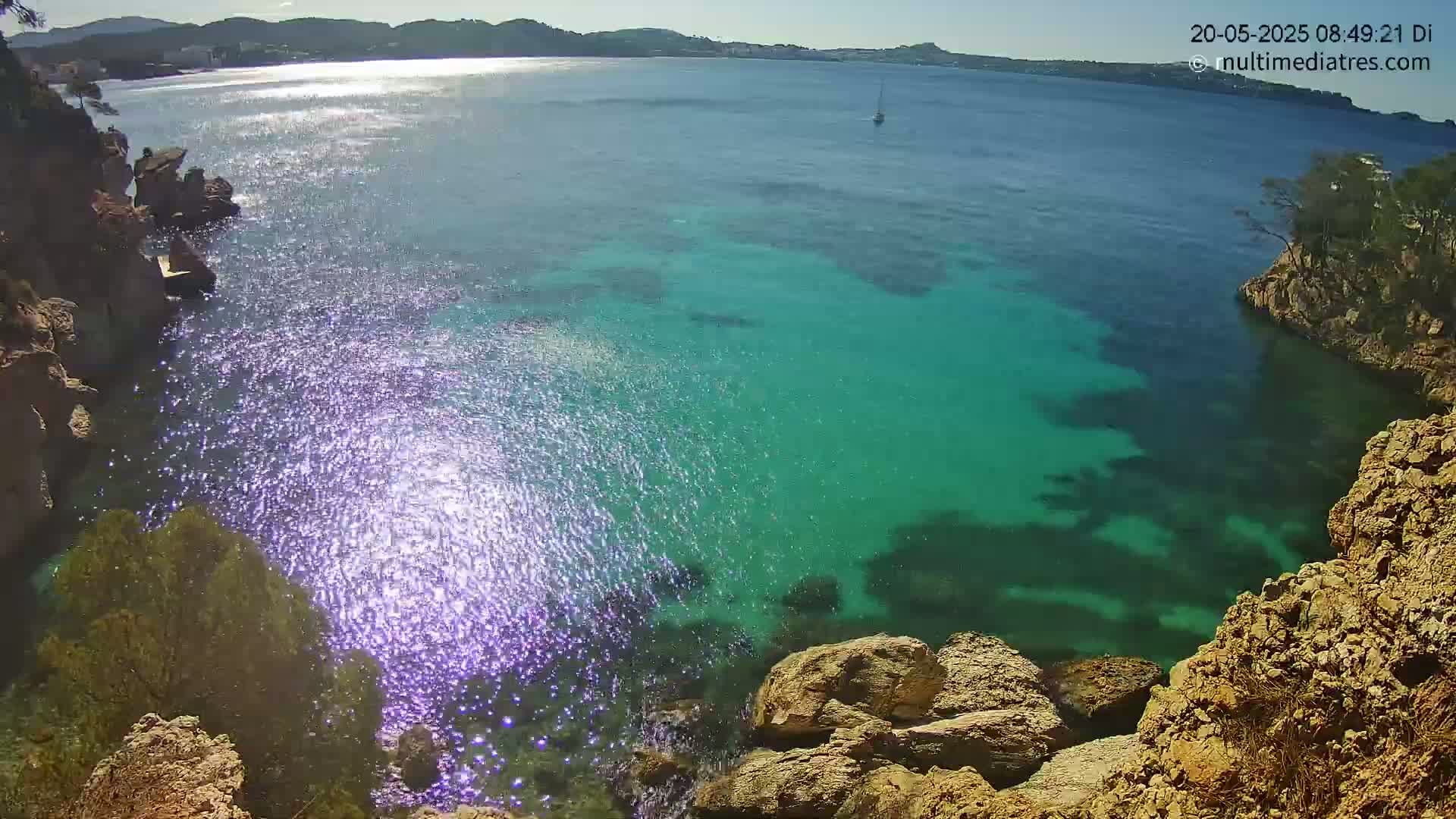 A high-angle view shows a cove of bright turquoise water sparkling in the sunlight, surrounded by rocky, tan-colored cliffs and greenery, with a sailboat visible in the distance on a sunny day.