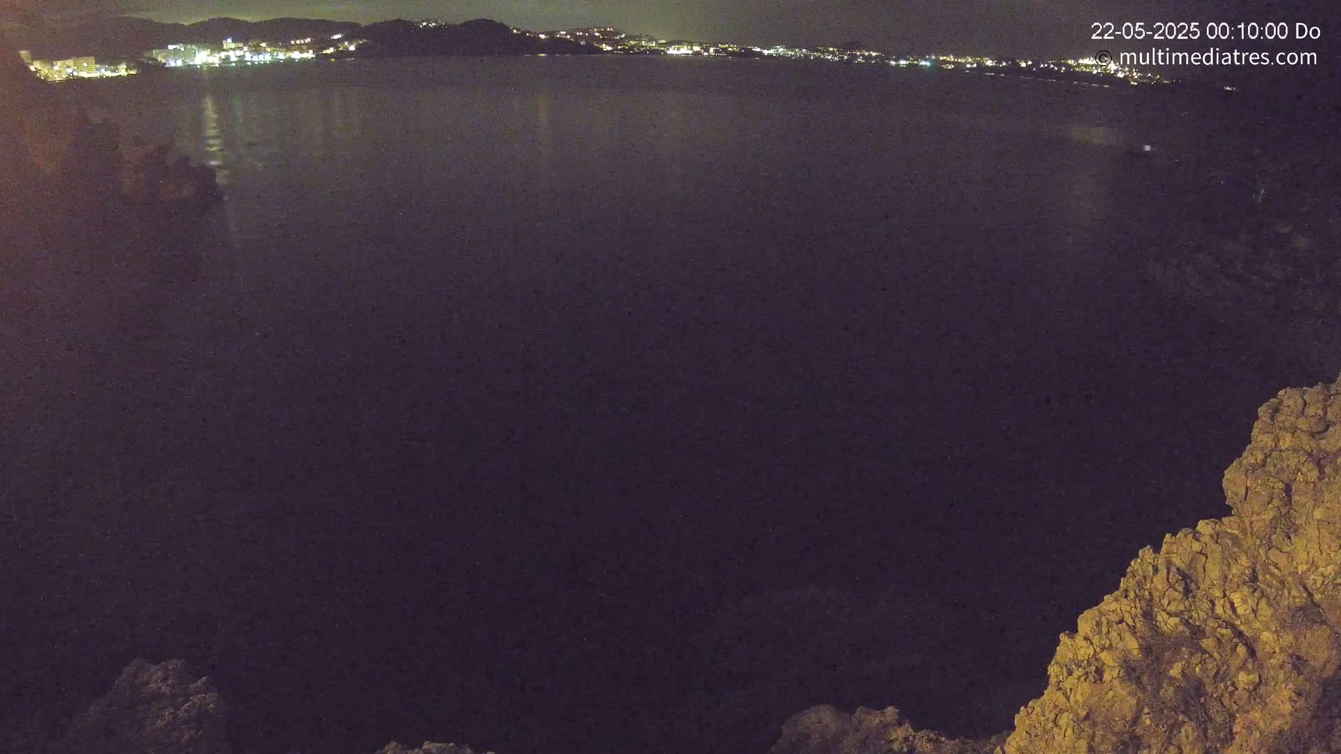 A nighttime view shows a dark body of water reflecting the lights of a distant town, framed by dark, rocky terrain.