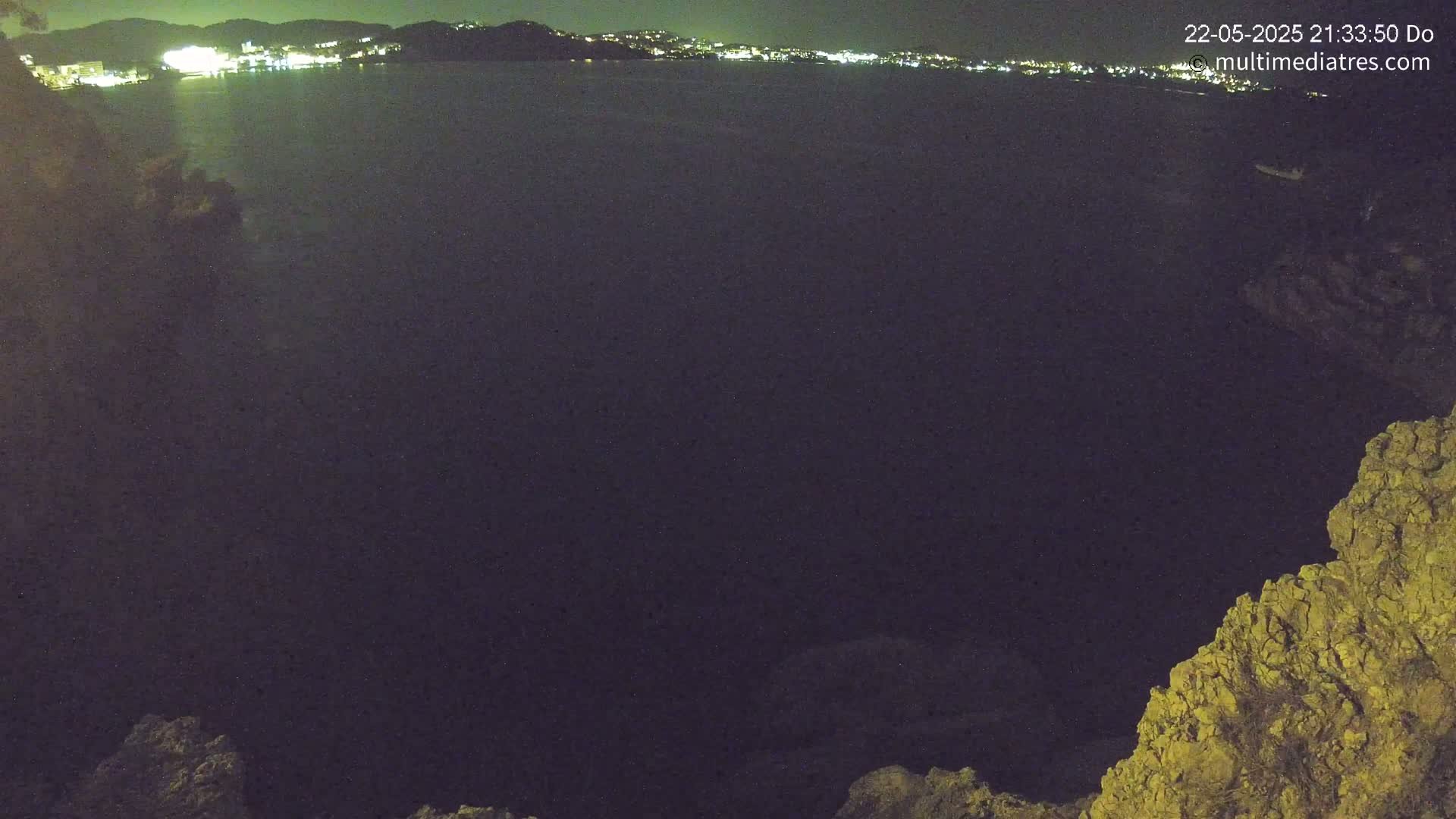 A nighttime view of a dark body of water, with a distant, illuminated shoreline visible in the background and rocky terrain in the foreground.