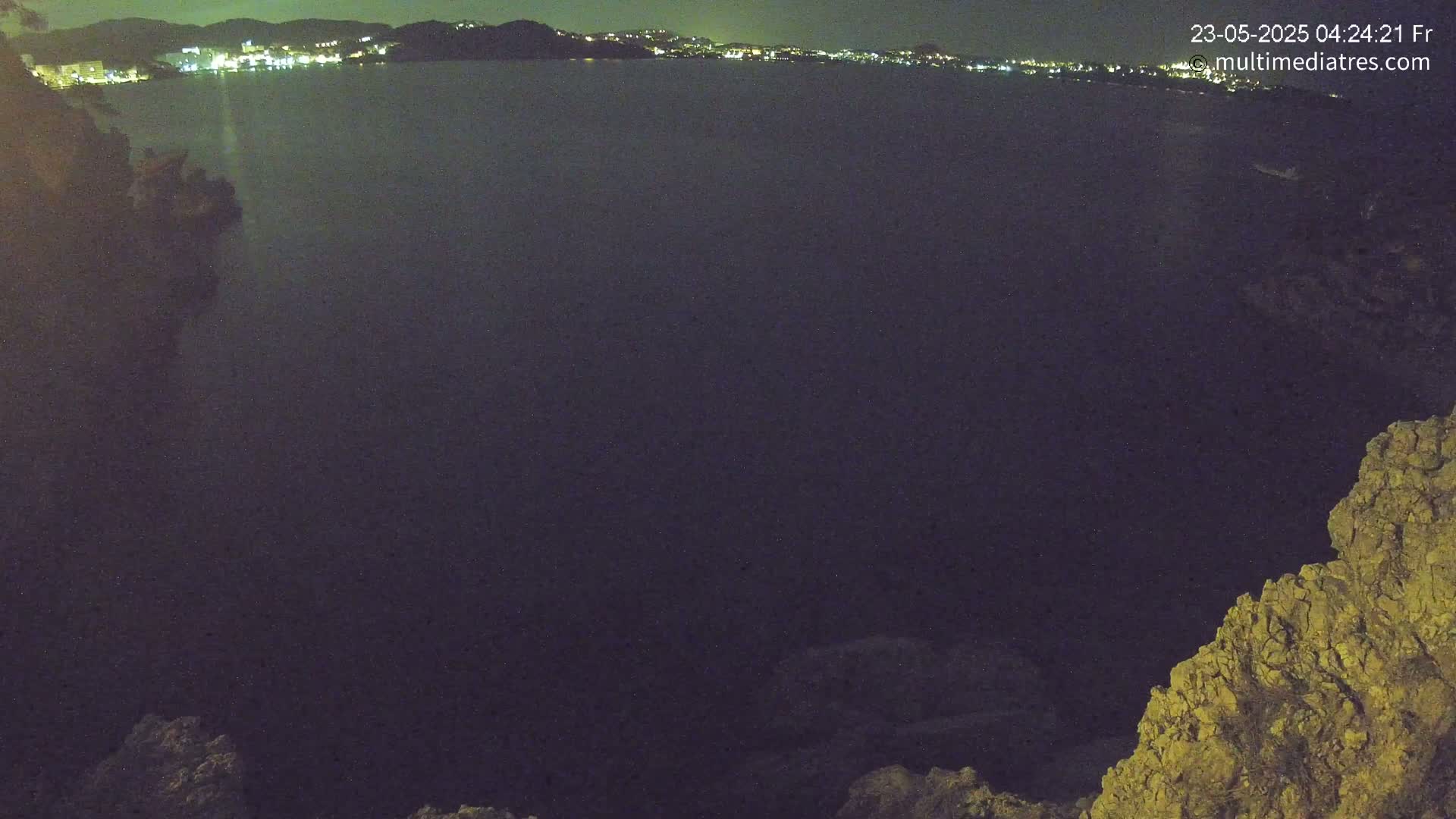 A nighttime view of a dark body of water with a distant, illuminated town visible on the horizon, partially obscured by a rocky outcrop in the foreground.