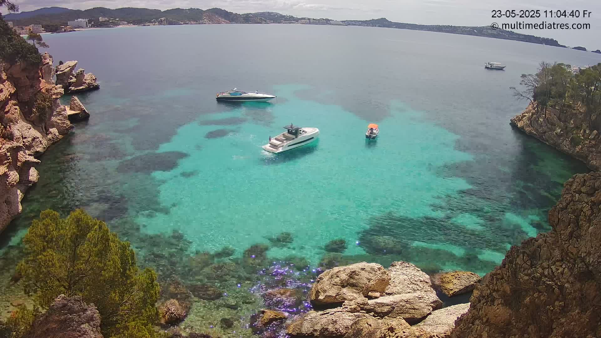 Three boats are anchored in a calm, clear turquoise cove surrounded by rocky, tan cliffs under a partly cloudy sky.