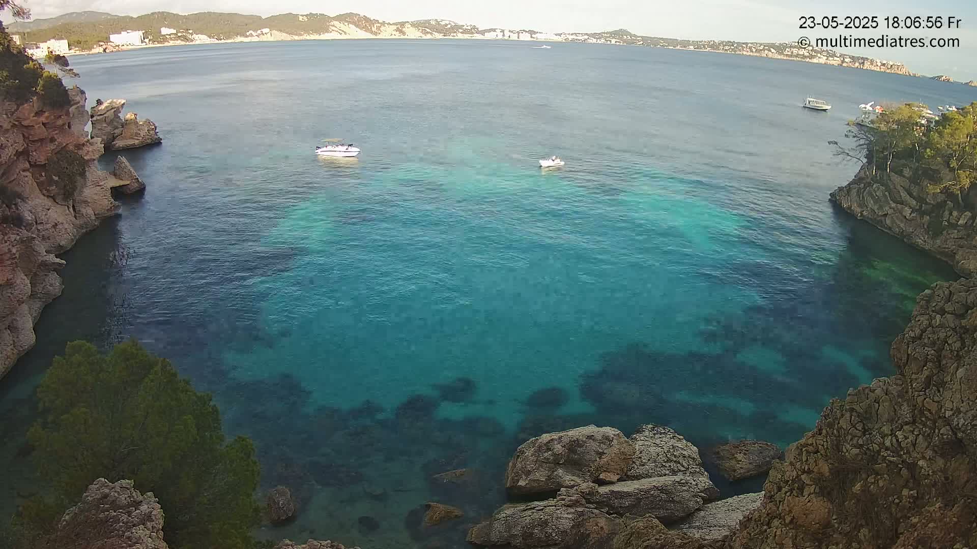 A calm, clear turquoise cove is nestled between rocky cliffs, with a few small boats visible in the distance under a sunny sky.