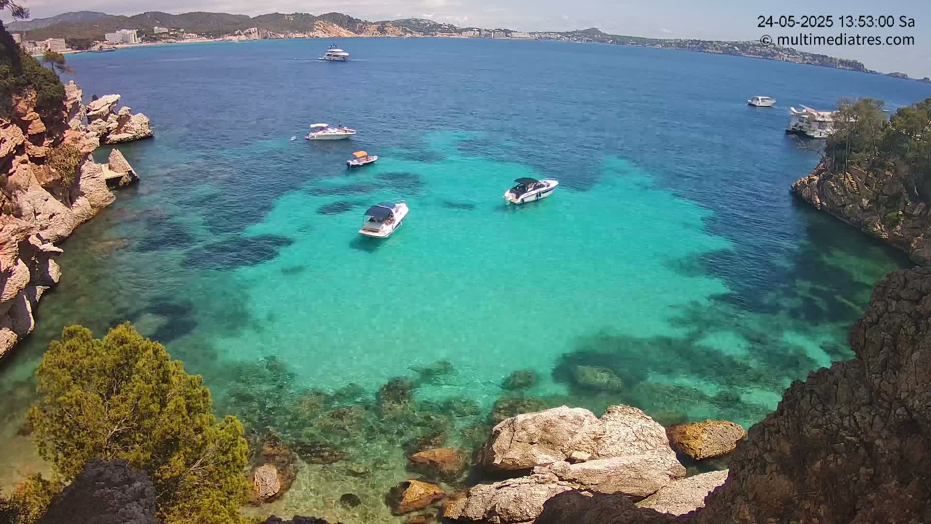 Several boats are anchored in a calm, clear turquoise cove surrounded by rocky cliffs and vegetation under a sunny sky.