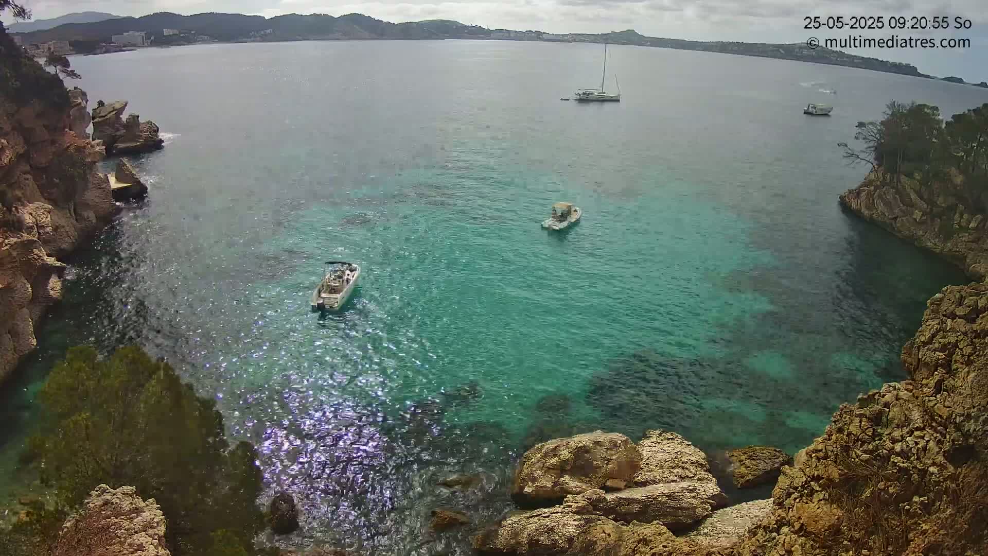 A tranquil cove with clear, turquoise water is nestled between rocky cliffs, with several small boats visible on the water under a partly cloudy sky.
