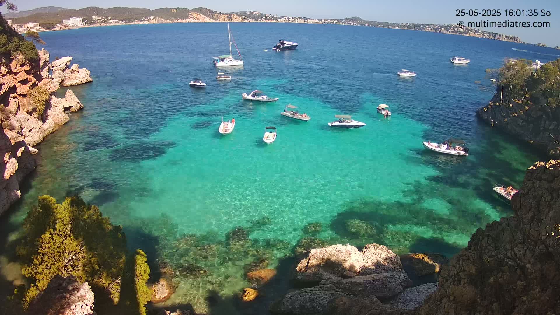 A calm, sunny day reveals a cove of clear, turquoise water surrounded by rocky cliffs, with several small boats and yachts scattered across the surface.
