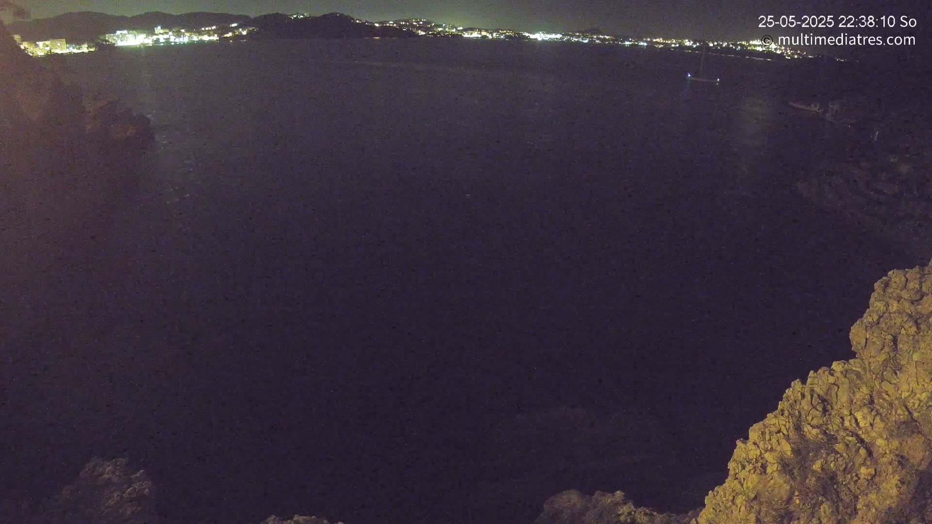 A nighttime view of dark water, a distant town's lights, and rocky terrain in the foreground.