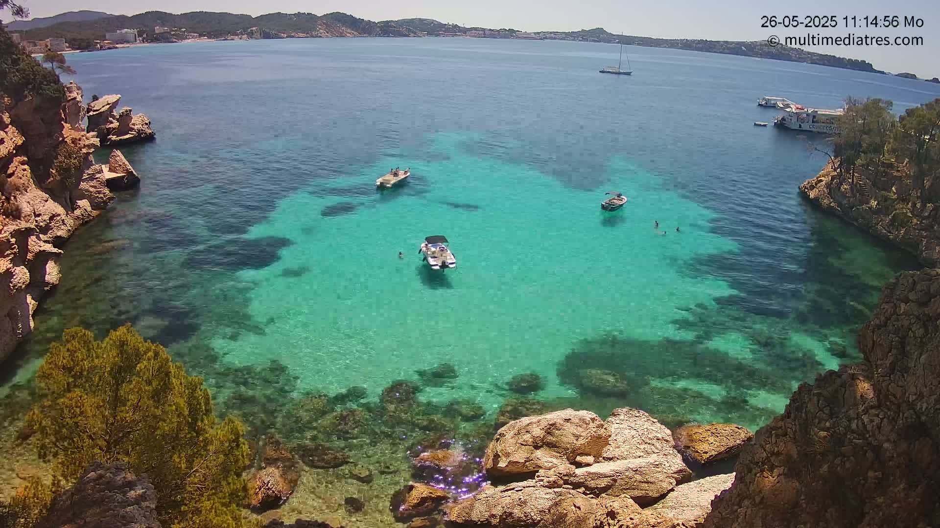 A tranquil cove with clear turquoise water, surrounded by rocky cliffs and several small boats, under a sunny sky.