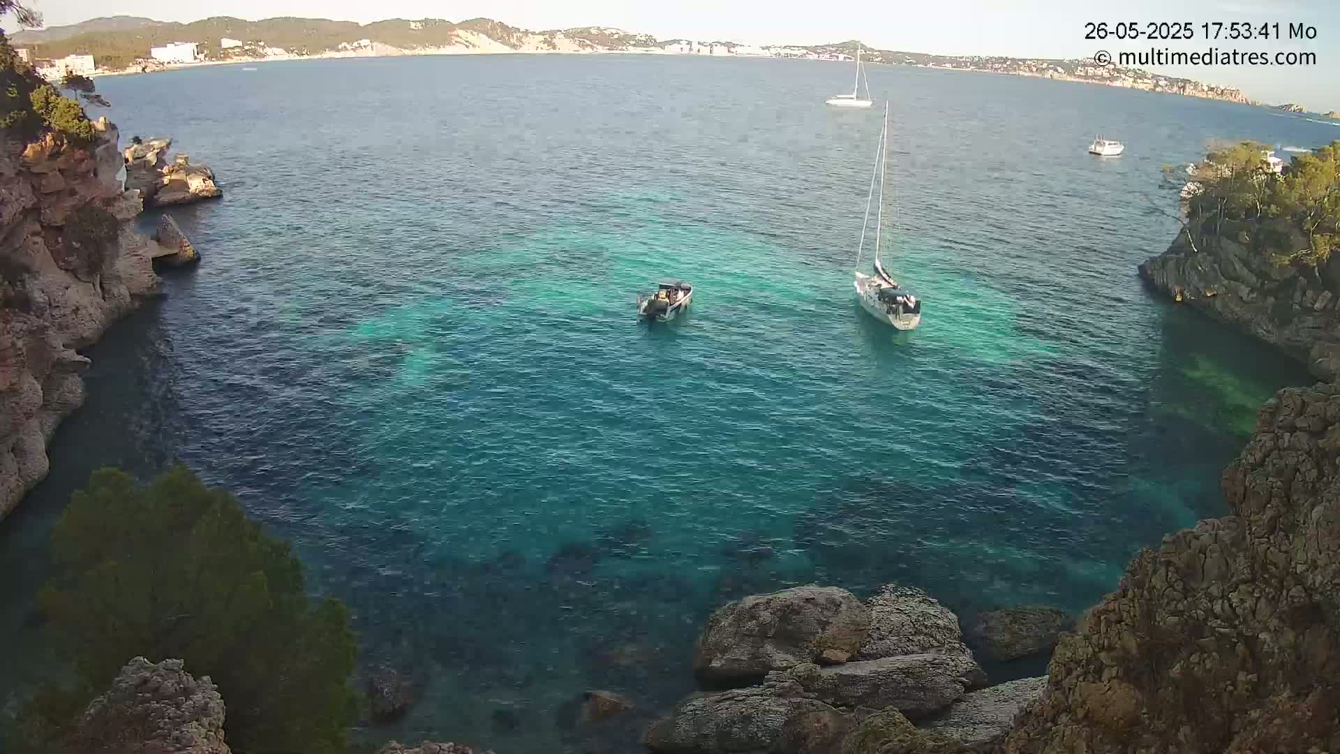 A tranquil cove of clear turquoise water is nestled between rocky cliffs, with a sailboat and a smaller motorboat visible in the calm sea.
