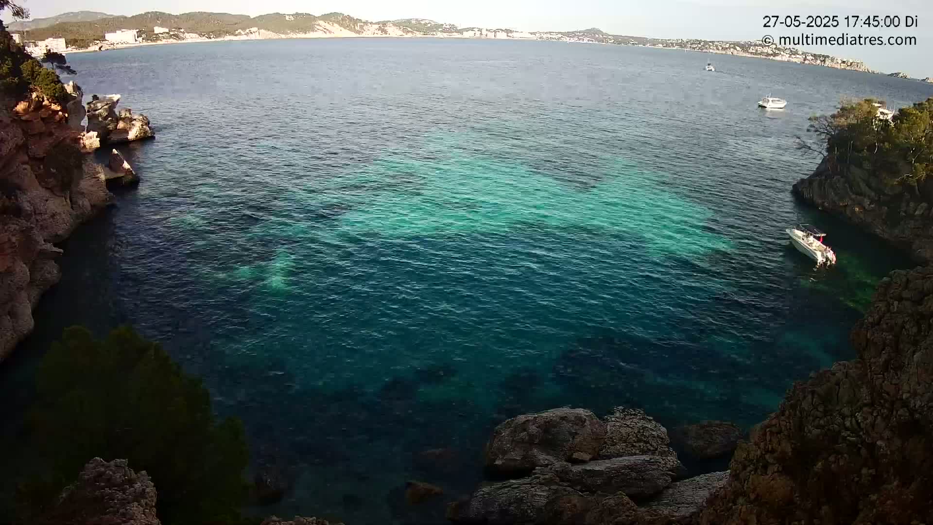 A clear, calm body of teal water is nestled between rocky shores, with a few boats visible in the distance and one closer to the shore.