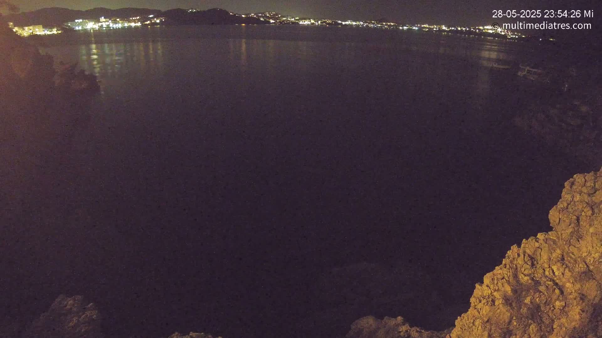 A nighttime view of a calm body of water reflecting the lights of a distant town, seen from a rocky vantage point.