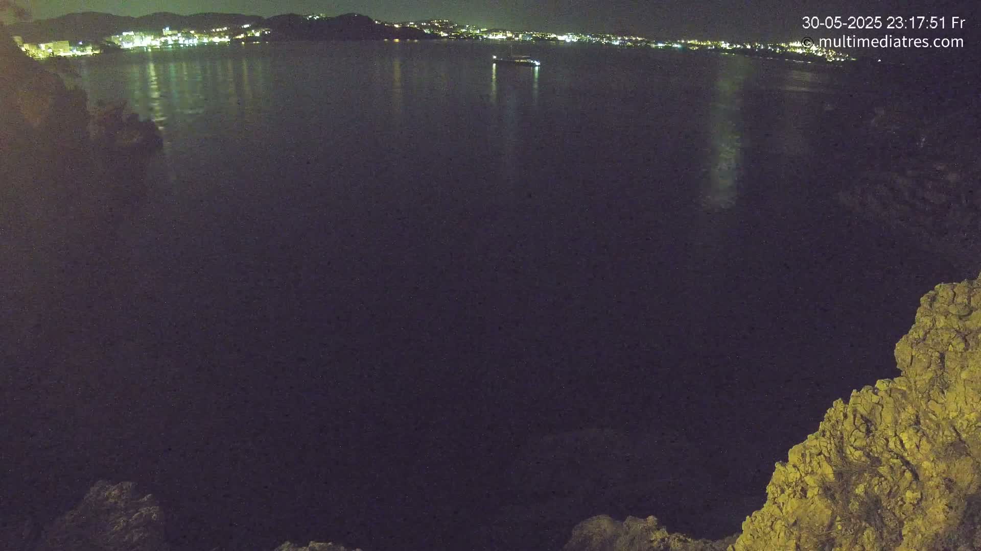 A nighttime view shows a dark body of water reflecting the lights of a distant town on a calm, clear night.