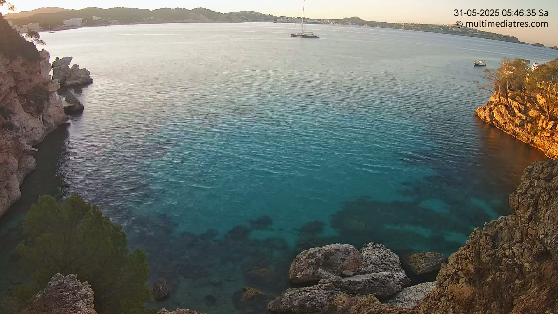 A tranquil cove of clear, teal water is nestled between rocky, tan cliffs under a calm, sunrise sky.