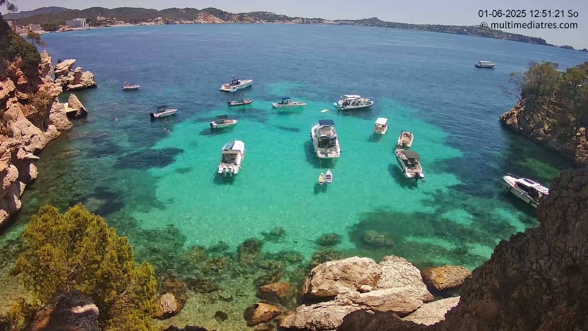 A sunny day shows a secluded cove with crystal-clear turquoise water, surrounded by rocky cliffs and numerous boats anchored in the calm sea.