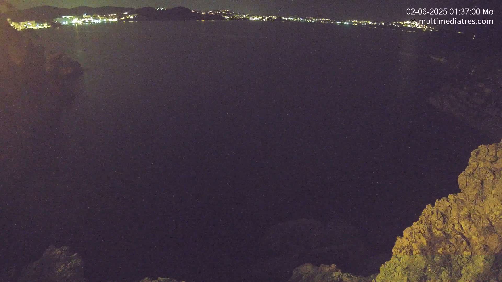 A nighttime view of a dark body of water with a distant, illuminated town visible across the water, partially obscured by a rocky outcrop in the foreground.