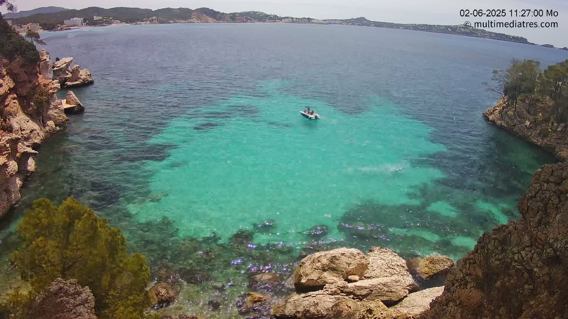 A small boat sits in a cove of clear, turquoise water surrounded by rocky cliffs and vegetation under a sunny sky.