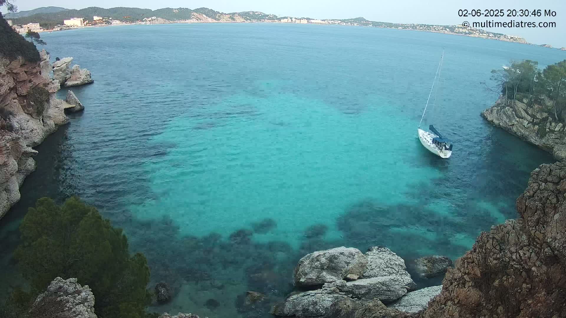 A sailboat is anchored in a calm, turquoise cove surrounded by rocky cliffs and overlooking a distant coastline.
