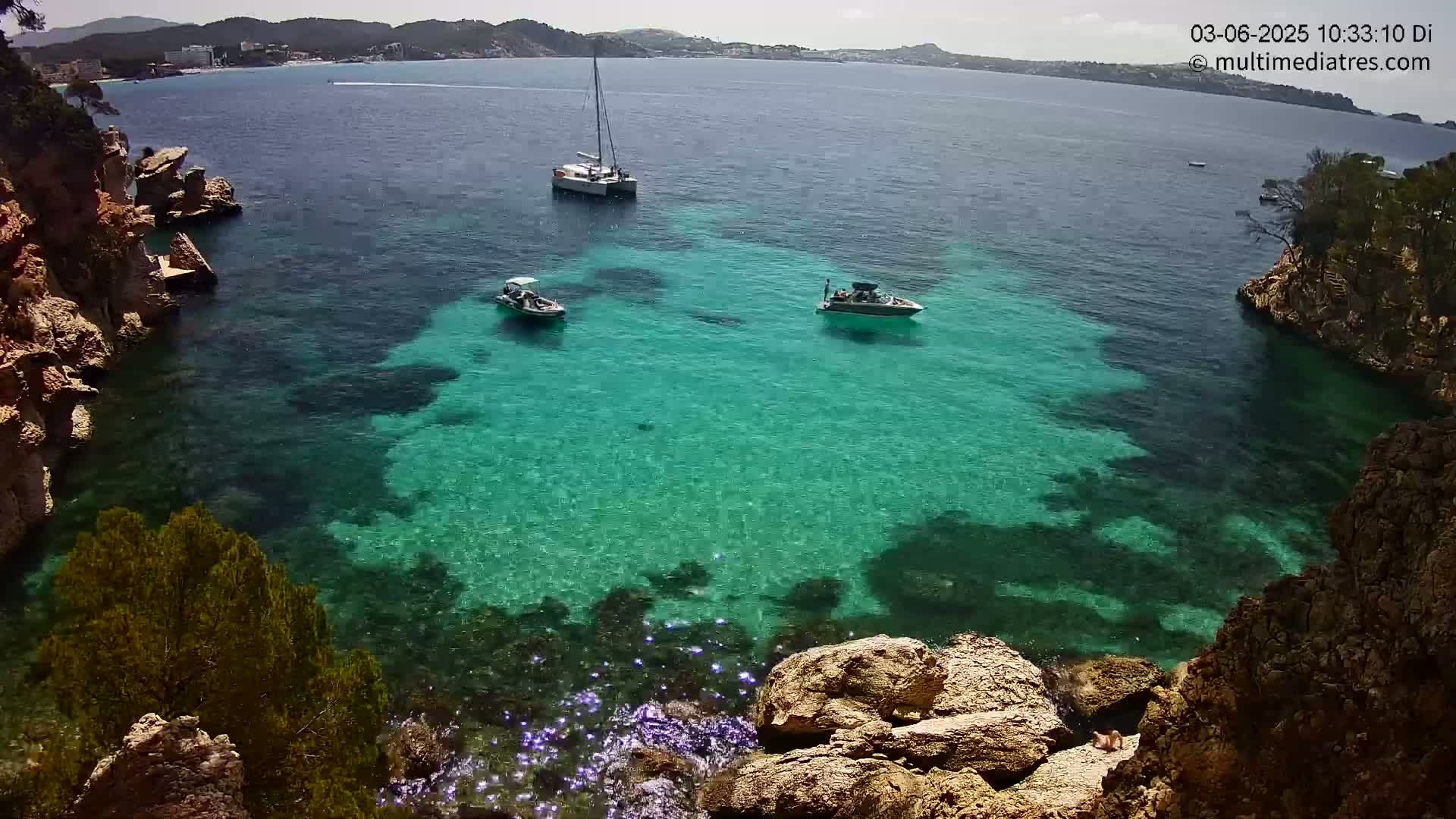 A secluded cove with crystal-clear turquoise water, surrounded by rocky cliffs and vegetation, contains a sailboat and two motorboats on a sunny day.