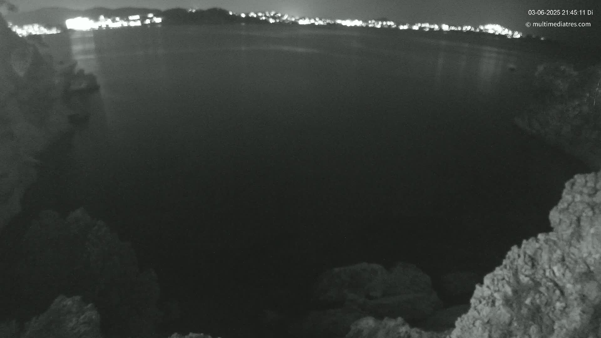 A nighttime view of a dark body of water, with a distant city skyline faintly illuminated, is seen from a rocky vantage point.