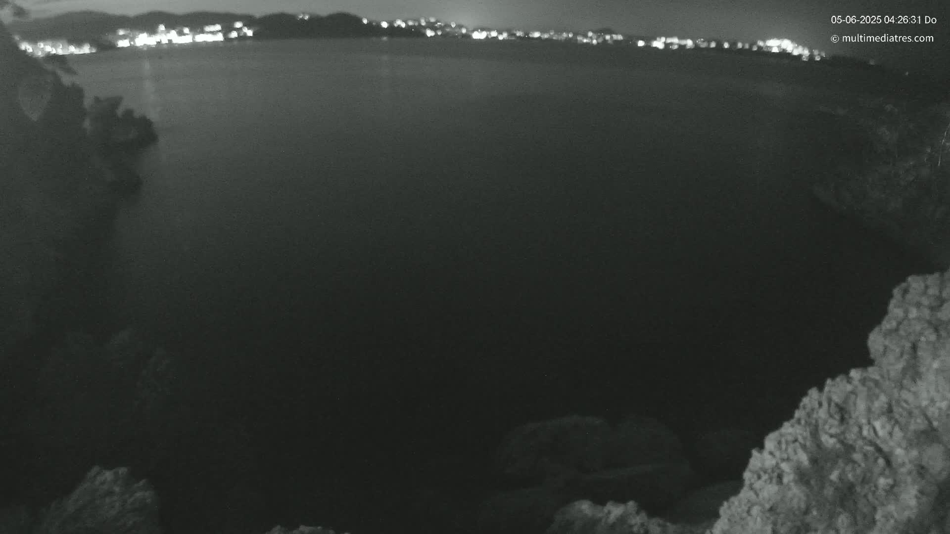 A nighttime view shows a dark body of water with a distant city skyline faintly illuminated, framed by rocky, uneven terrain.