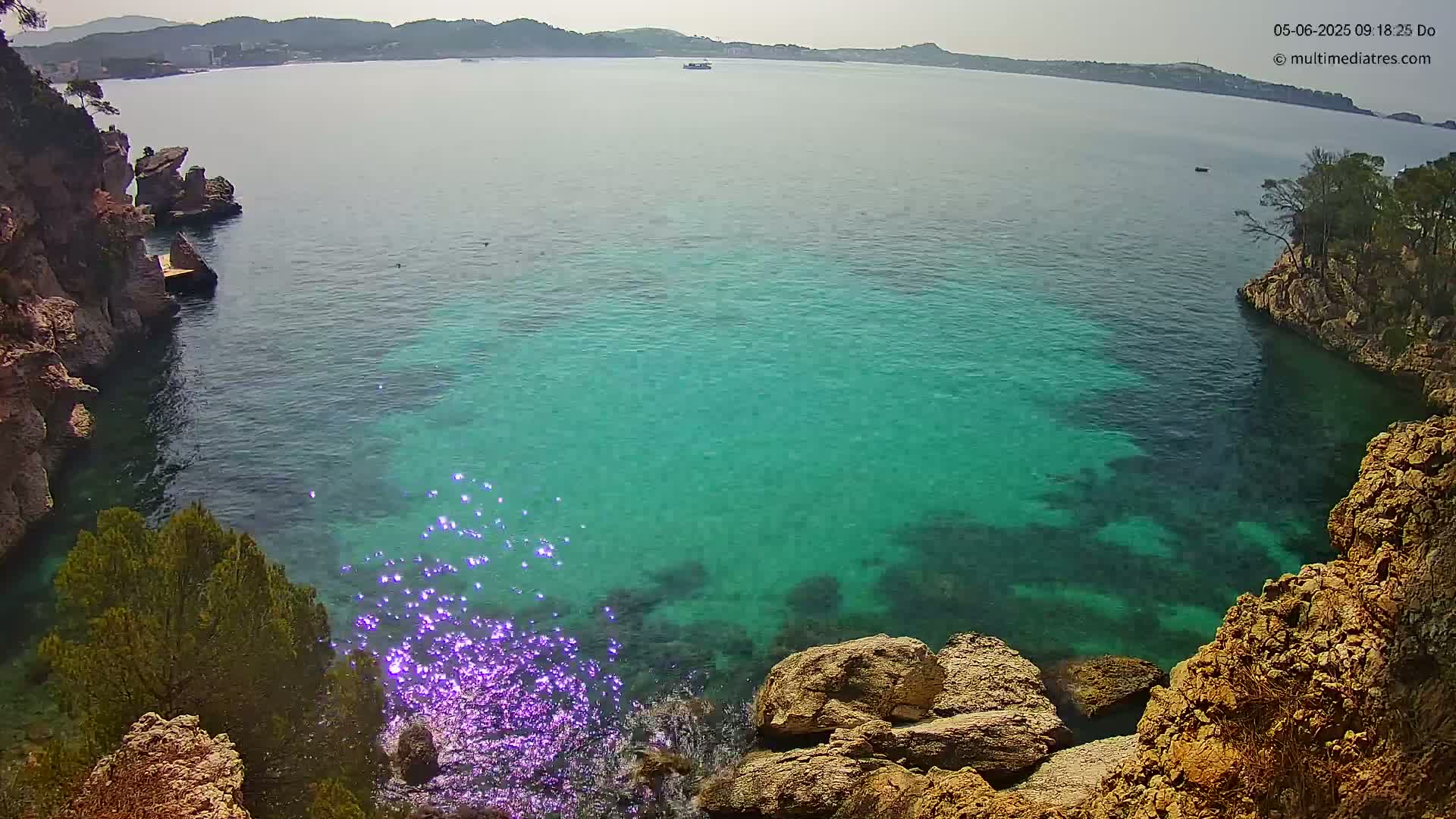 A cove of clear, turquoise water is nestled between rocky, vegetation-covered cliffs on a sunny day.