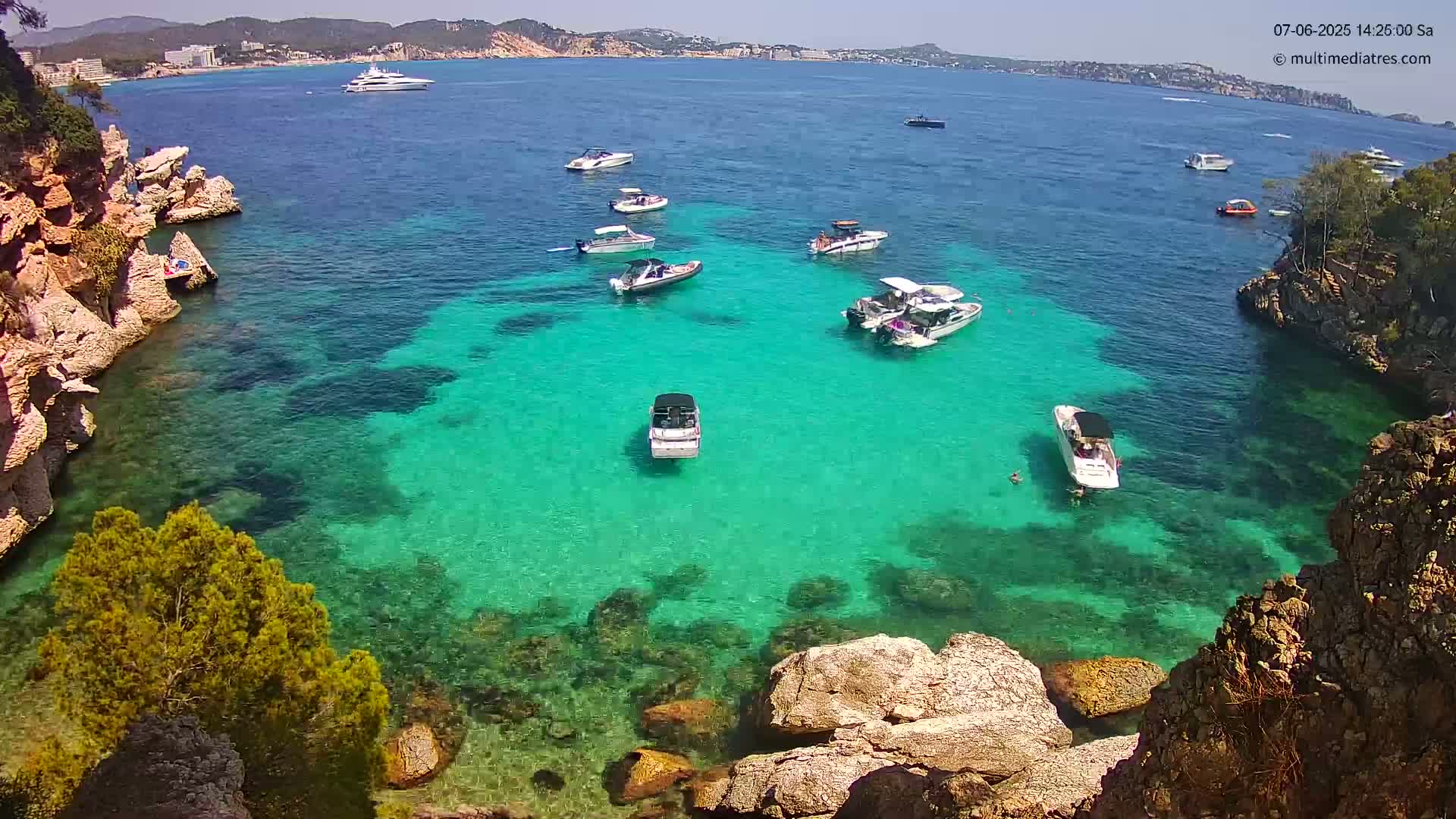A cove with crystal-clear turquoise water is surrounded by rocky cliffs and several small motorboats are anchored in the calm, sunny water.