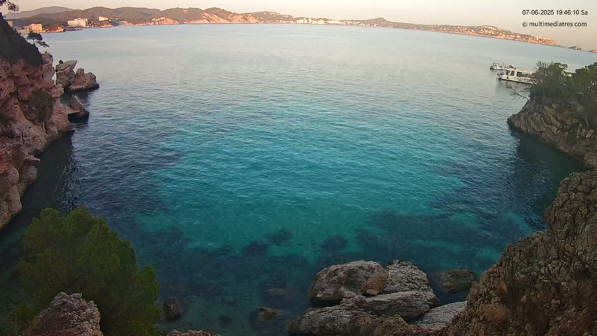 A tranquil cove of clear, teal water is nestled between rocky cliffs, with a distant coastline visible under a calm evening sky.
