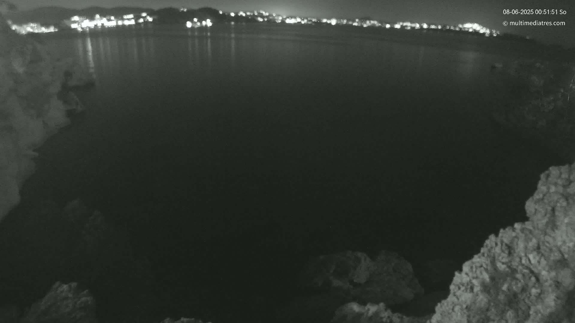 A nighttime, dark, and calm body of water reflects the lights of a distant town, framed by rocky terrain.