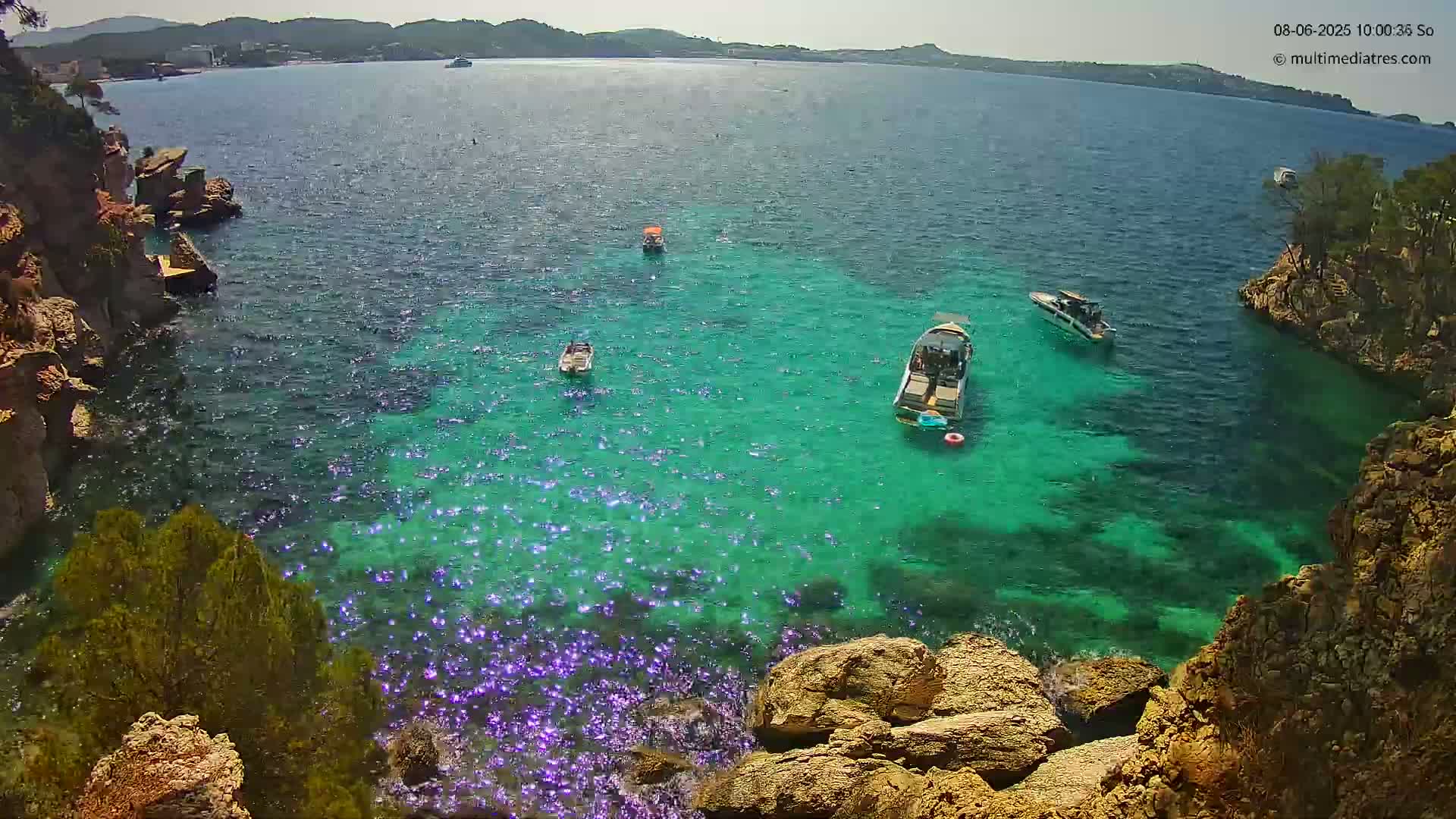 Several boats are anchored in a calm, clear, bright-green cove surrounded by rocky, Mediterranean-style cliffs on a sunny day.