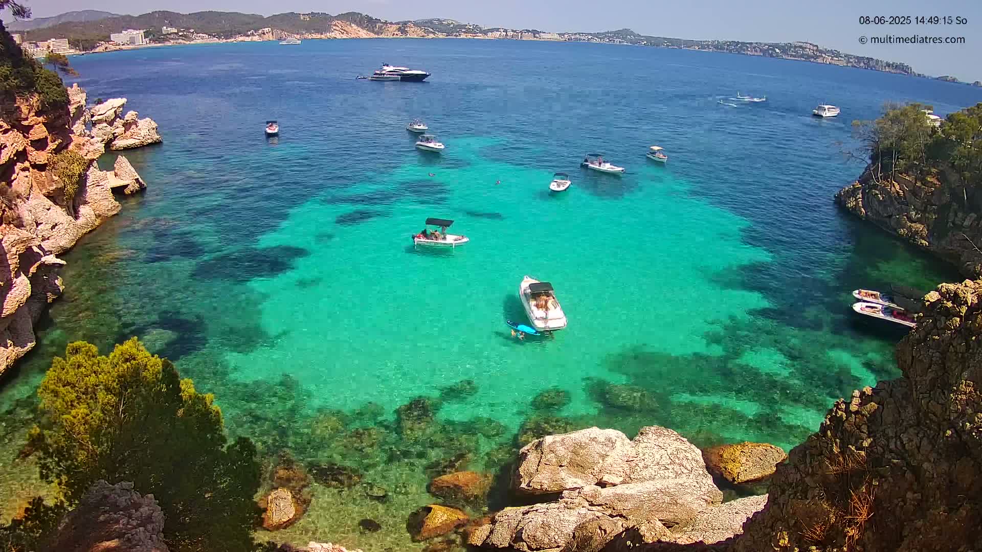 Clear turquoise water filled with several small boats is surrounded by rocky, tan-colored cliffs under a sunny sky.