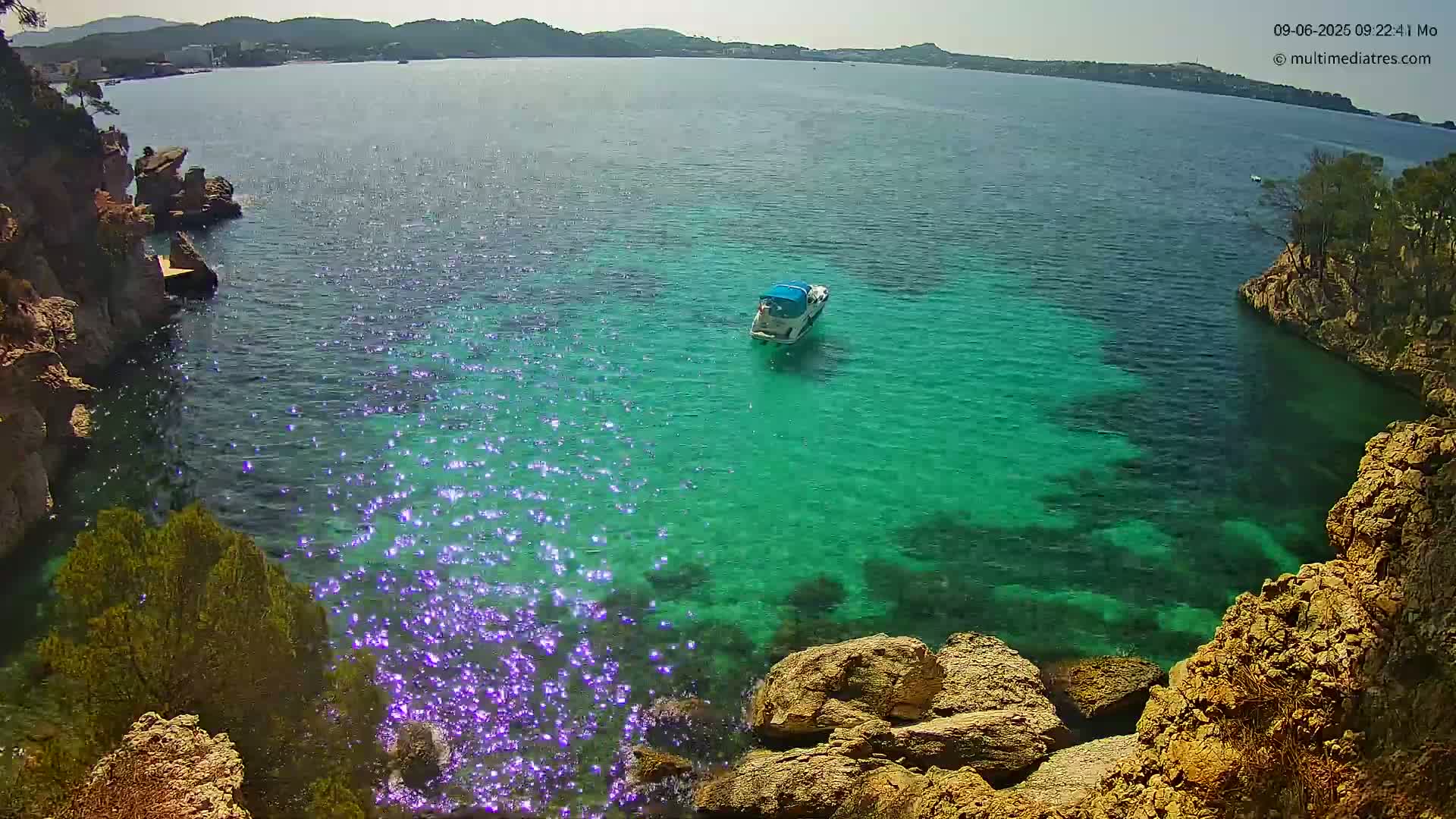 A small motorboat sits in a calm, sparkling, teal cove surrounded by rocky, vegetated shores on a sunny day.