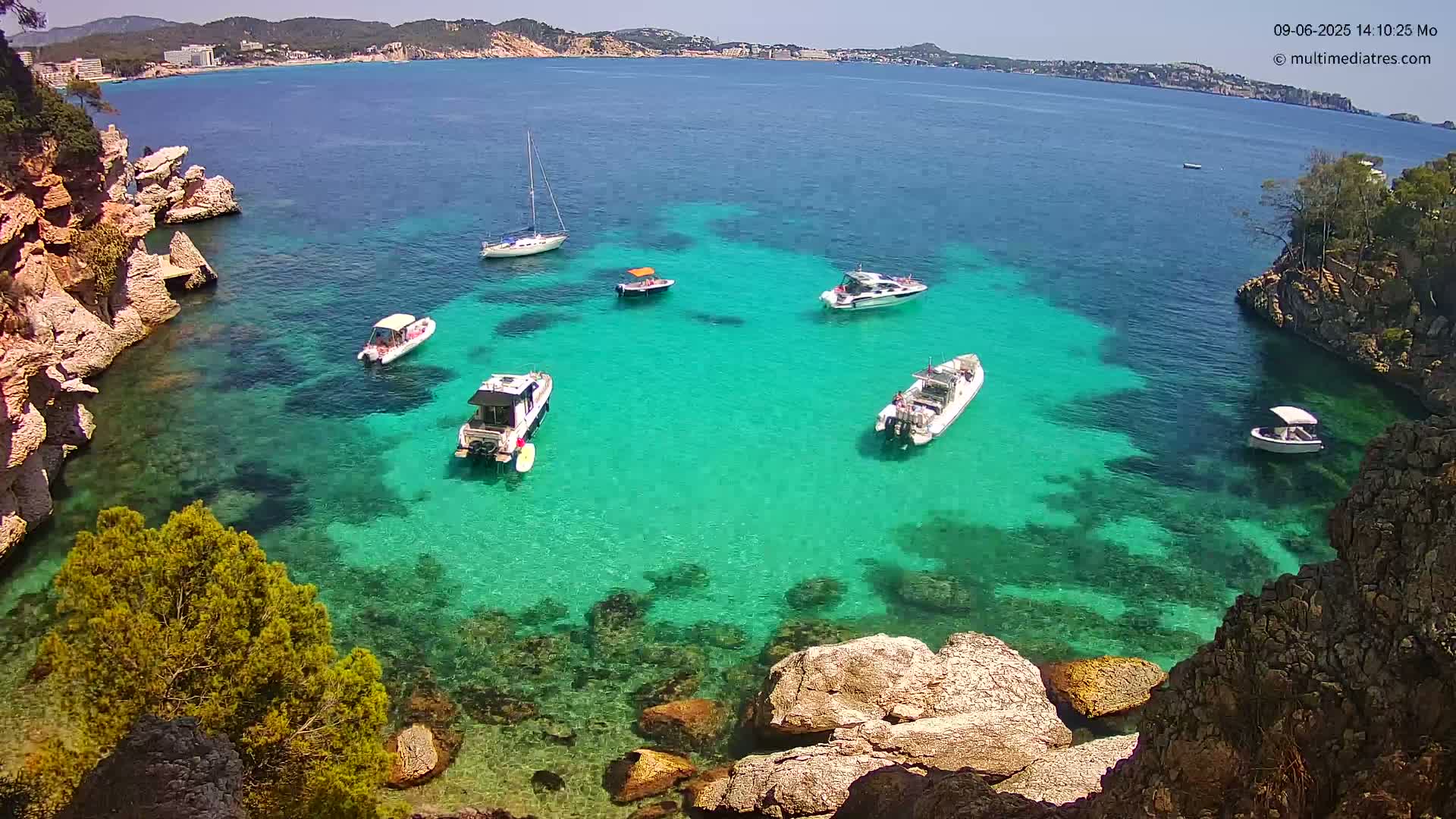 A tranquil cove with clear, turquoise water is surrounded by rocky cliffs and contains several boats on a sunny day.