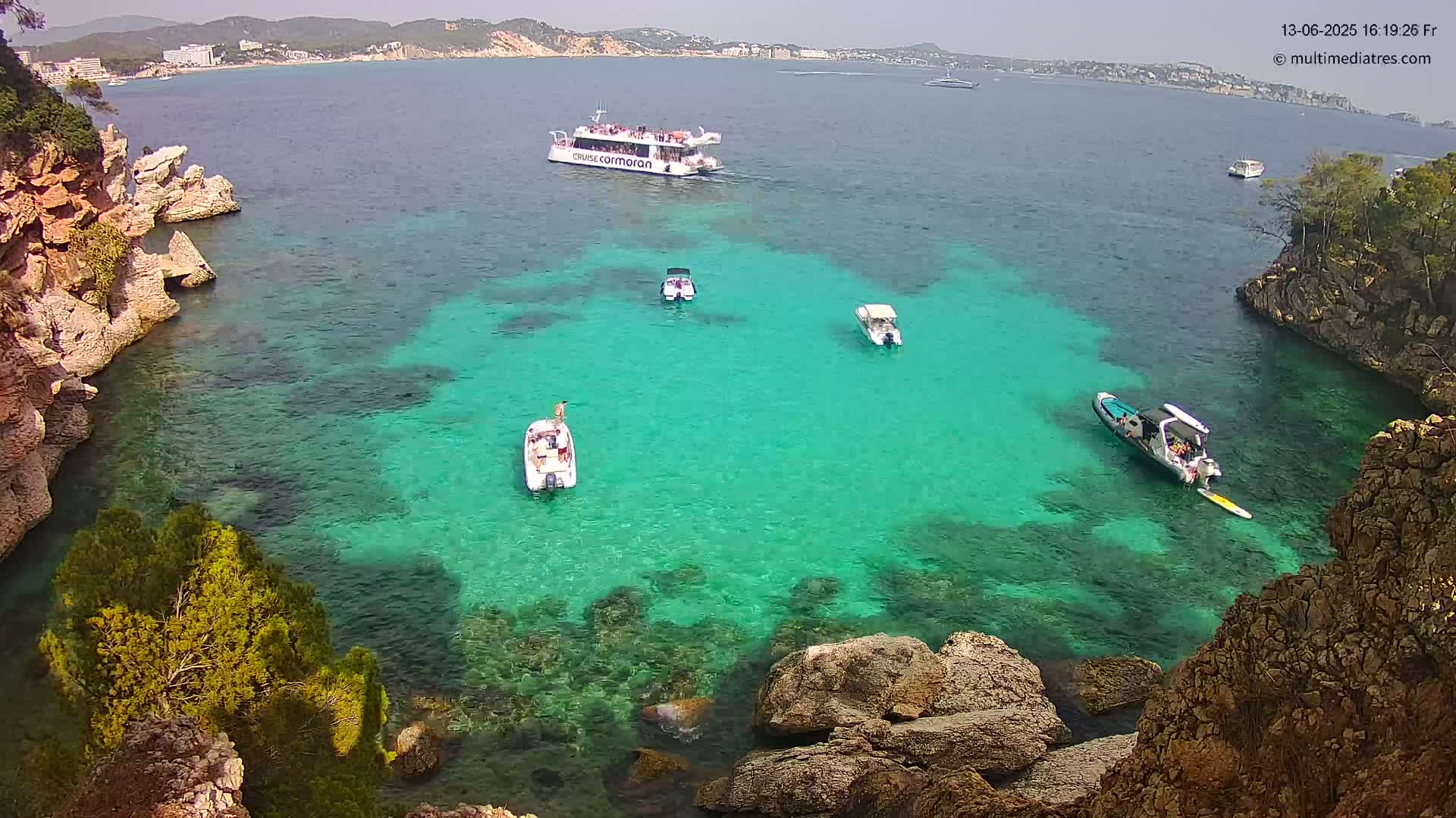 A bright, sunny day shows a secluded cove of clear turquoise water surrounded by rocky cliffs and several small boats, with a larger passenger boat visible in the distance.
