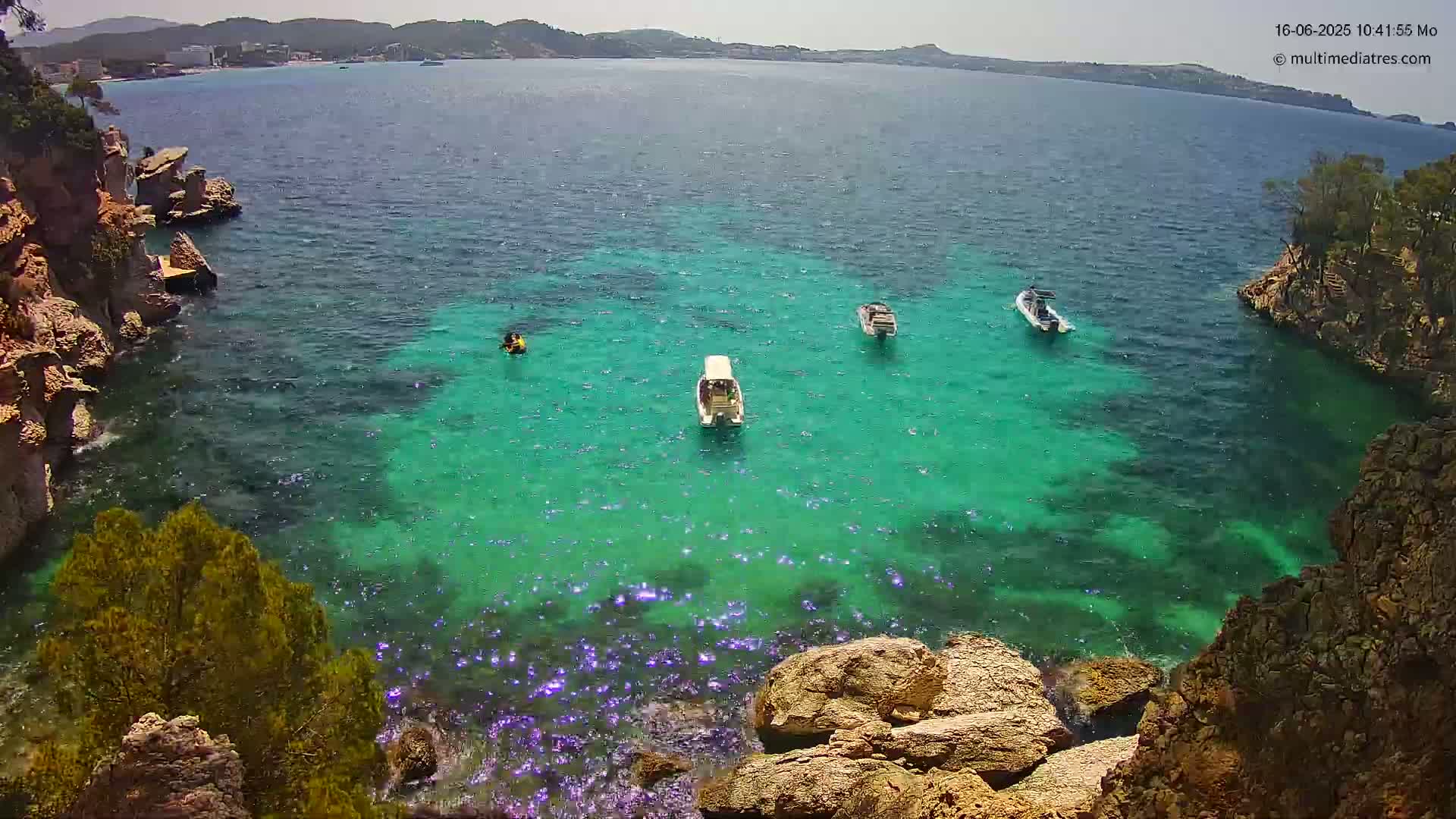 Clear, turquoise water surrounds rocky outcrops, with several small boats and a person on a jet ski visible on a sunny day.