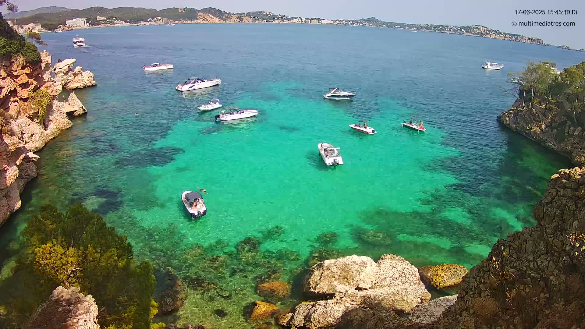 A tranquil cove with clear, turquoise water holds several small boats, surrounded by rocky, sunlit shores under a bright, sunny sky.