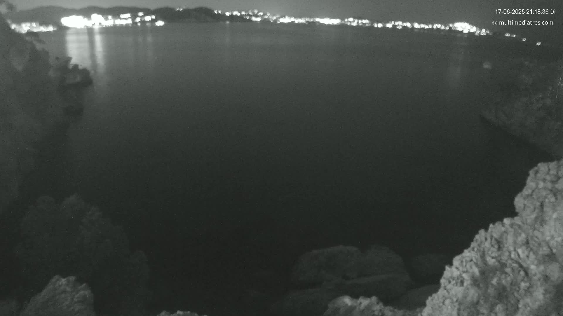 A nighttime, dark-grey body of water is seen from a rocky vantage point, with a distant city's lights faintly visible on the horizon.