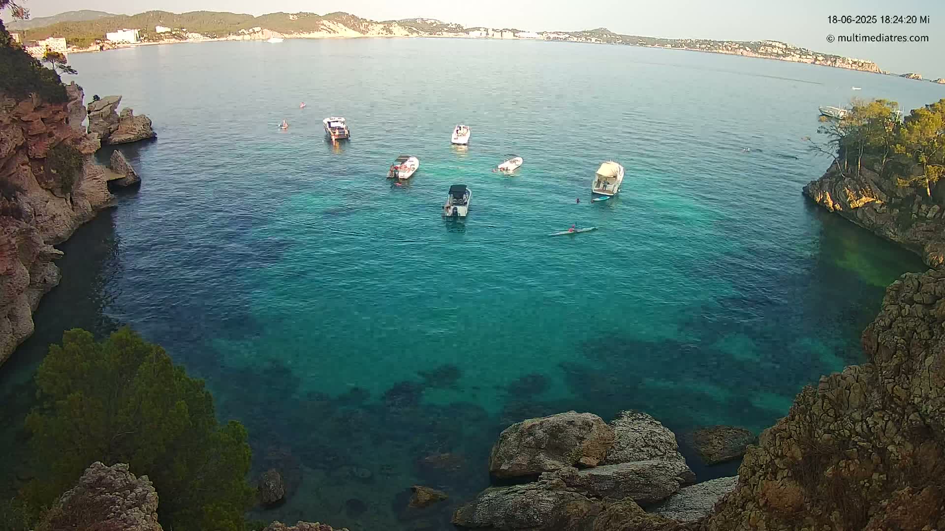 Several small boats are anchored in a calm, clear turquoise cove surrounded by rocky cliffs and vegetation.