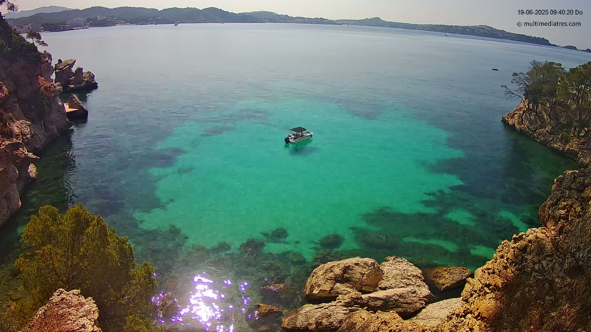 A small boat sits in a calm, bright teal cove surrounded by rocky cliffs and  trees on a sunny day.