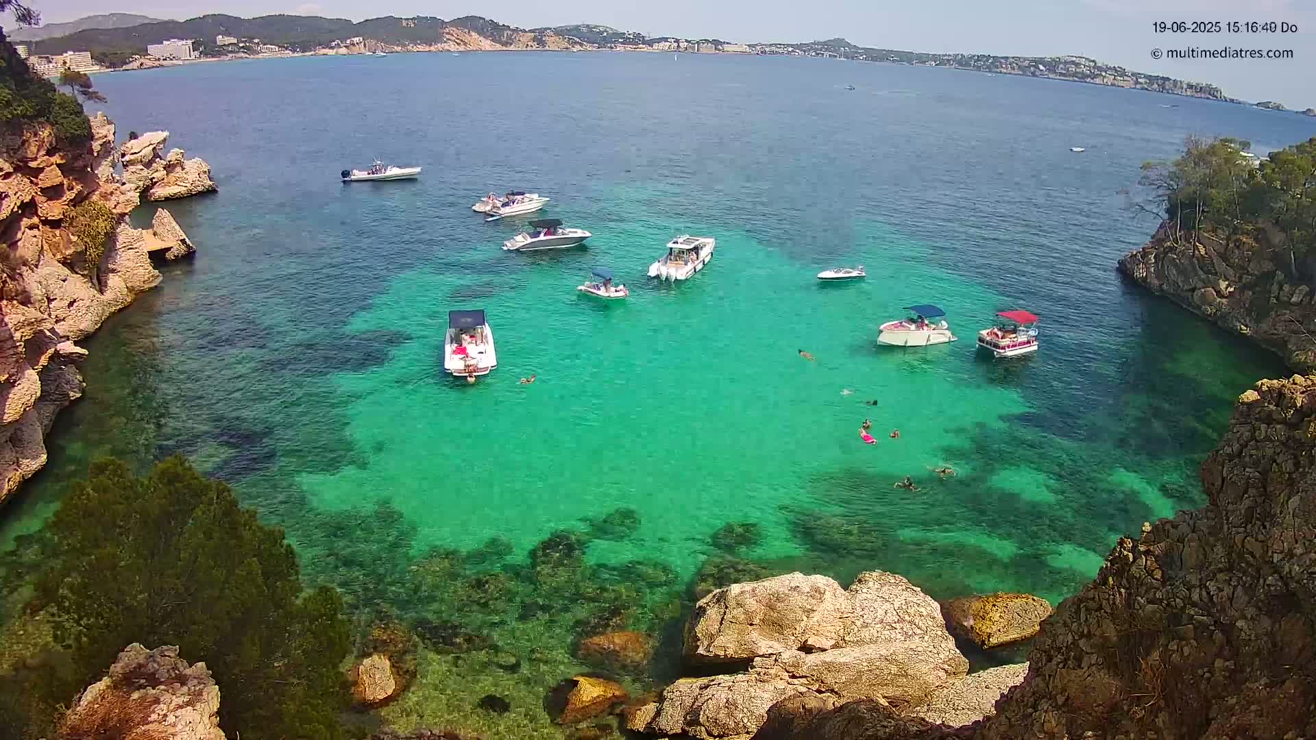 A cove with crystal-clear, teal water contains several boats and people swimming, surrounded by rocky, tan-colored cliffs on a sunny day.
