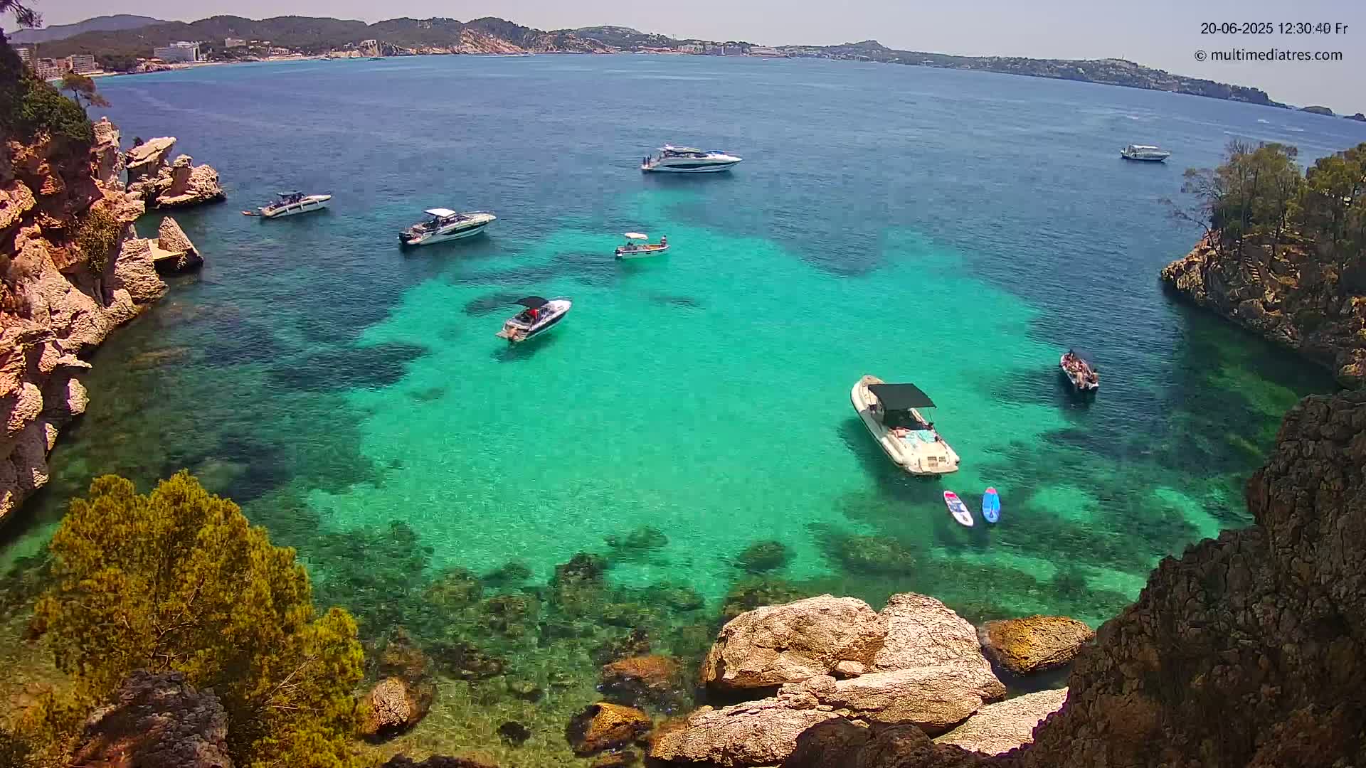 A cove of clear, teal water is surrounded by rocky cliffs and several boats are anchored in the calm, sunny weather.