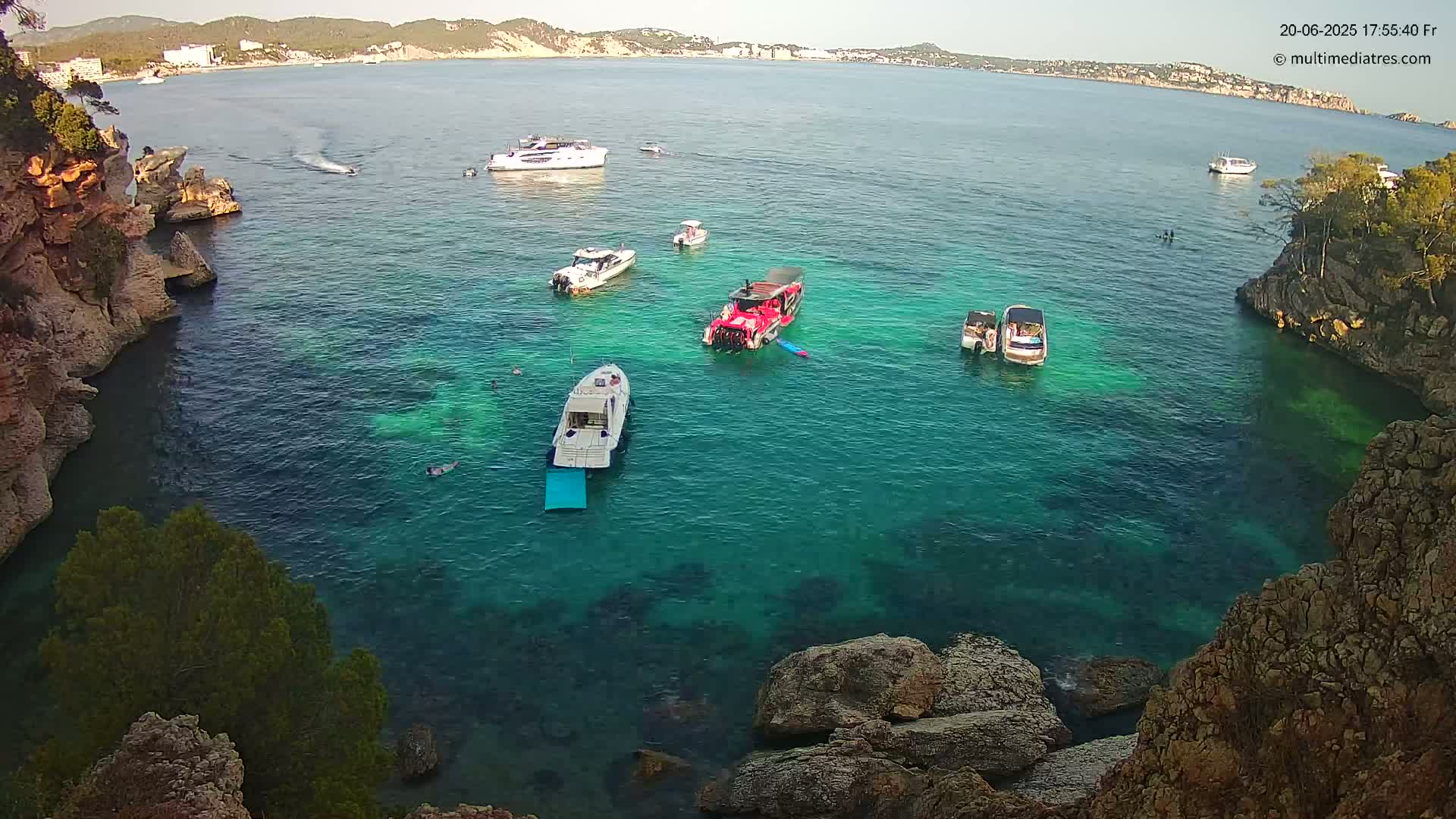 A calm, clear turquoise cove, surrounded by rocky cliffs and dotted with several boats, some carrying passengers.