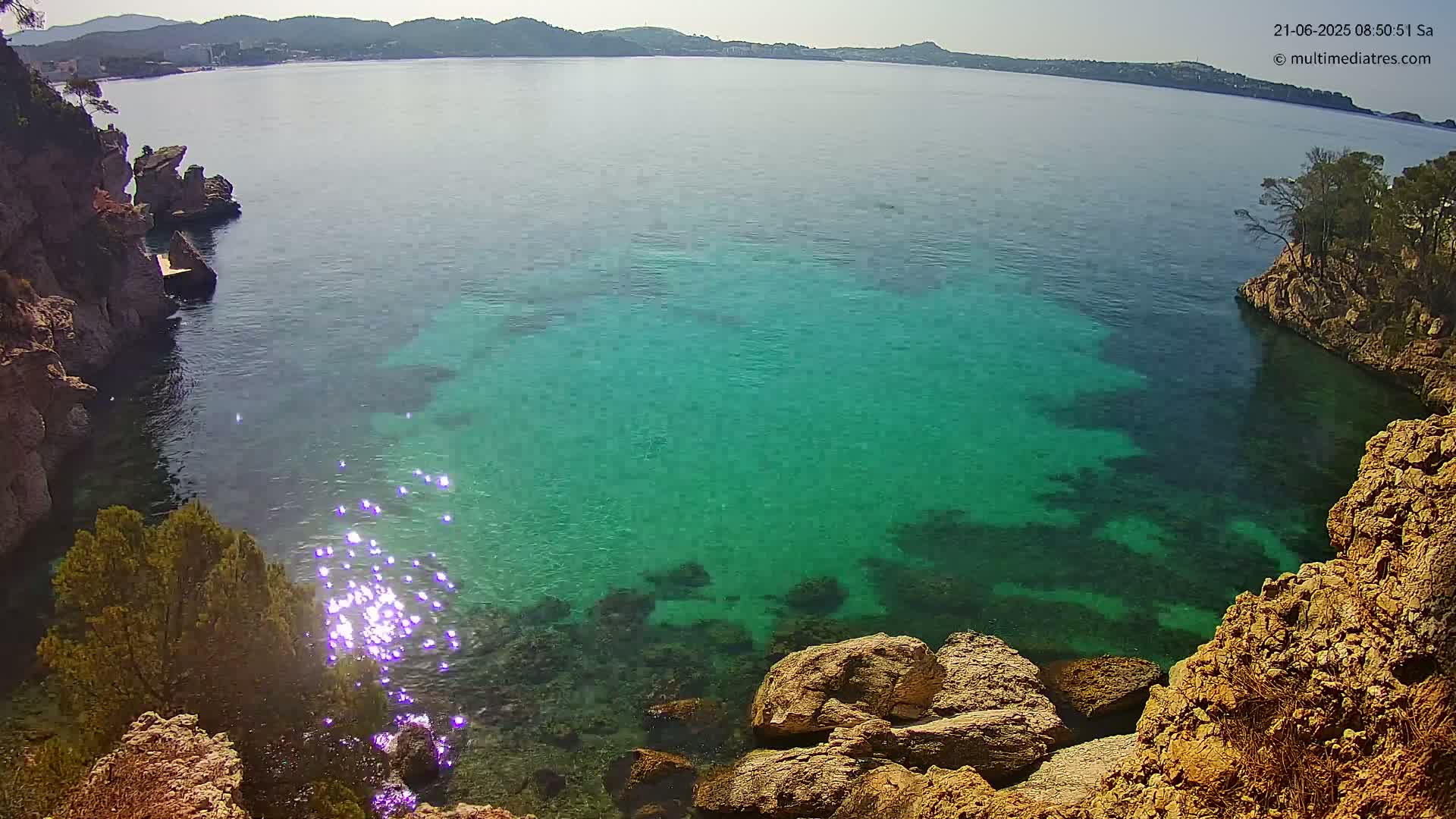 Bright sunlight glints off the clear, turquoise water of a secluded cove, surrounded by rocky, tan-colored cliffs and sparse green vegetation.