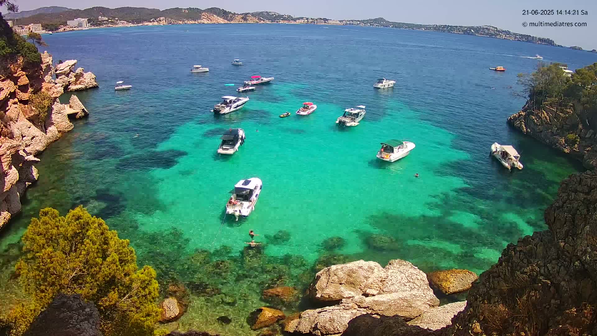 A tranquil cove with clear, turquoise water, numerous boats, and rocky shores under sunny skies.