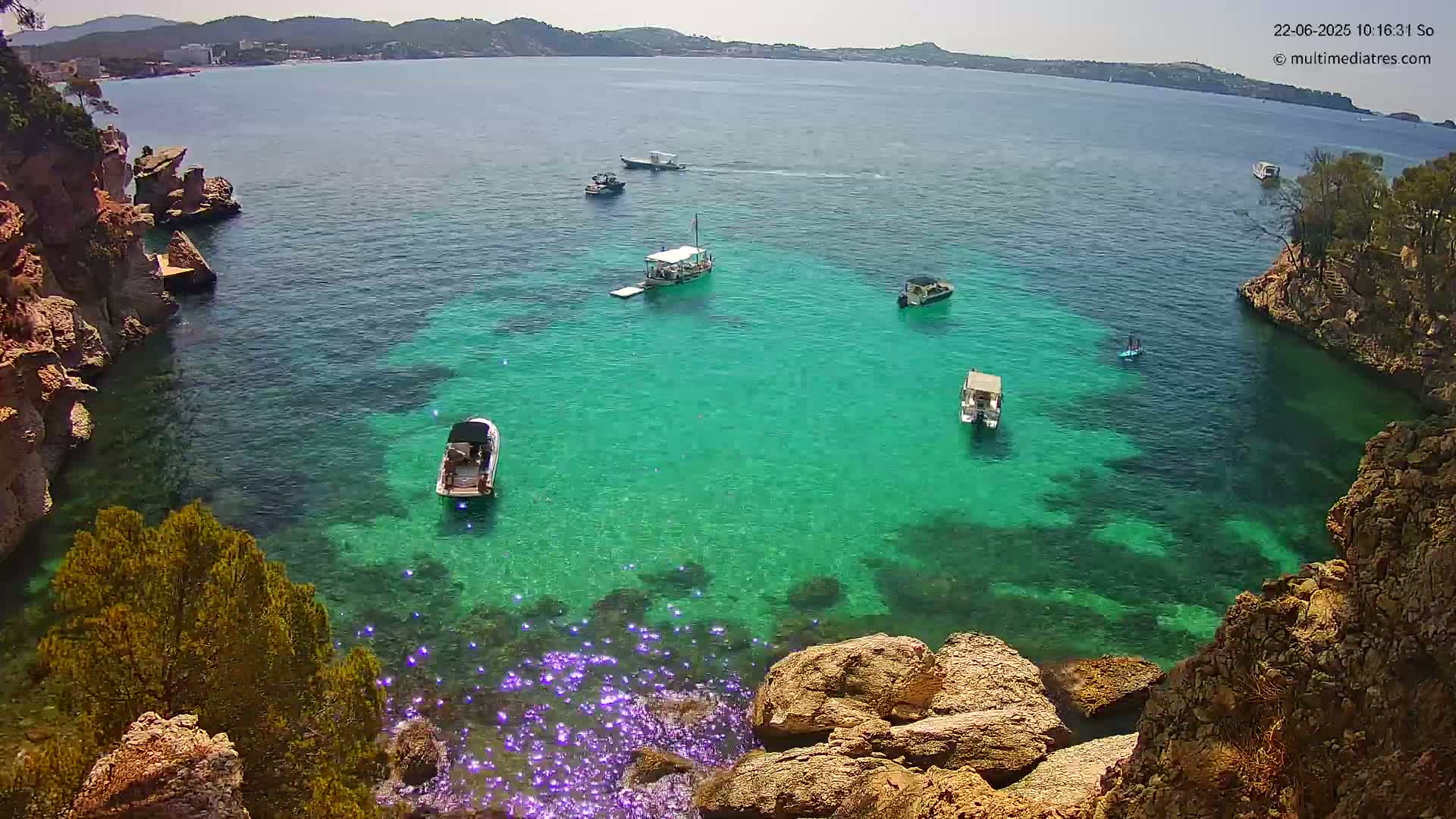 A cove of clear, bright teal water is surrounded by rocky, tan cliffs and several small boats are visible in the calm water.