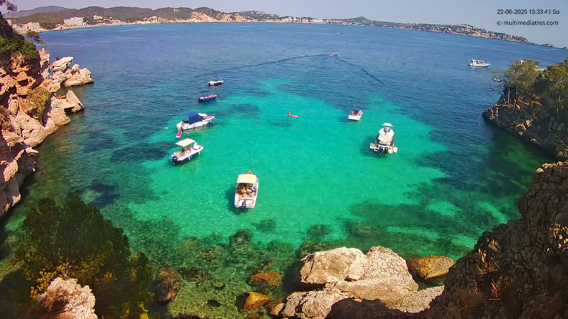 Several small boats are anchored in a calm, clear, bright teal cove surrounded by rocky cliffs under a sunny sky.