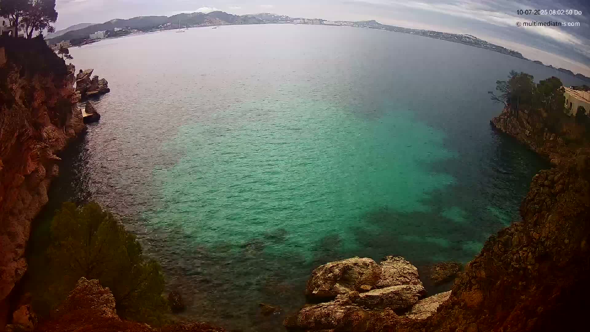 A high-angle view shows a calm, teal-colored bay nestled between rocky, vegetated cliffs under an overcast sky.