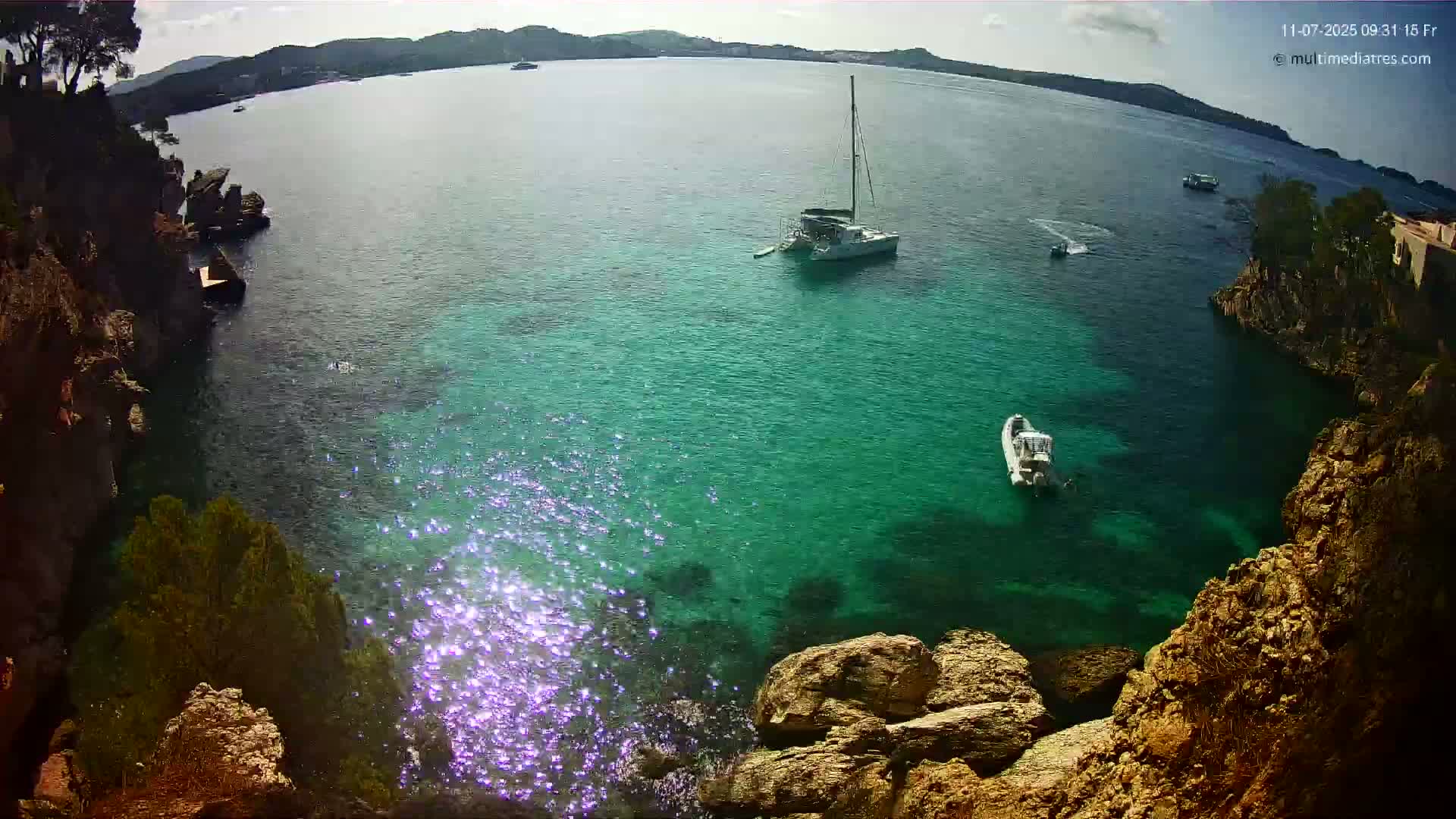 A bright, sunny day shows a secluded cove of clear, teal water with a sailboat and small motorboat anchored, surrounded by rocky cliffs and sparse vegetation.