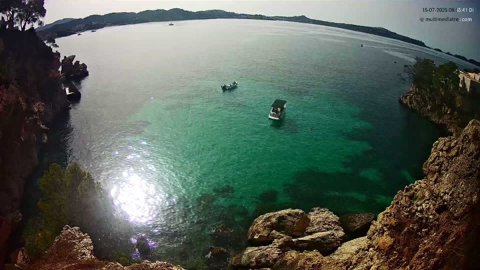 A bright, sunny day shows a cove of clear, teal water surrounded by rocky cliffs, with two small boats visible in the distance.